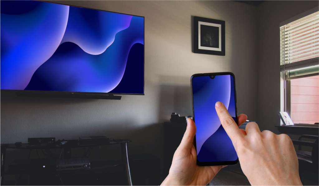 A hand holding an iPhone that is mirroring an image of blue waves to a wall-mounted Hitachi TV. The TV is located in a dimly lit living room
